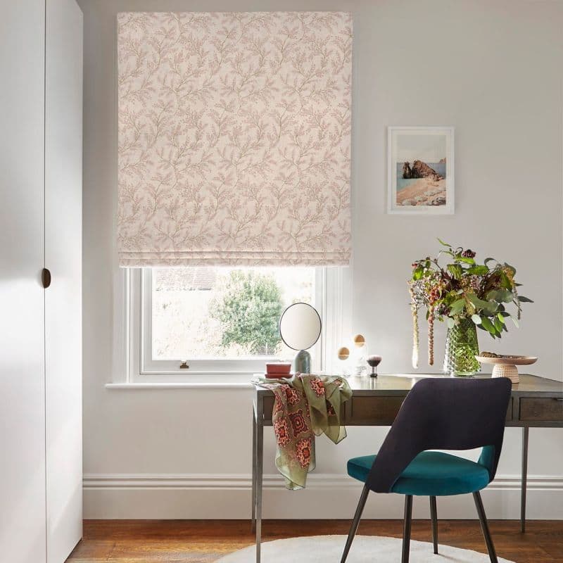 Floral-patterned Roman blind (pale pink, twig motif) lowered over the window, filtering daylight through a small bottom gap; above a desk with teal chair, vase of flowers, mirror, framed print.