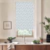 A patterned roller blind hangs partially lowered over a window above a kitchen sink with faucet, countertop, potted plants, a black bowl of greens, and a framed sketch.