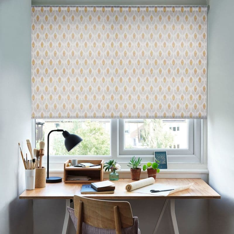 Patterned roller blind, lowered over the window—pale fabric with repeating yellow leaf motifs—filtering daylight; tidy wooden desk beneath with lamp, brushes, notebooks and small potted plants, urban tree view.