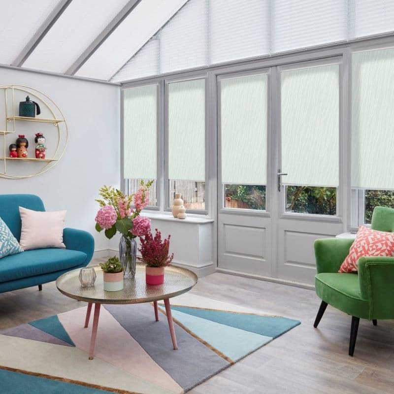 Pale mint, textured roller blinds lowered mid-height across tall conservatory doors and windows, diffusing daylight; cozy living room with teal sofa, green armchair, geometric rug, round coffee table with flowers.