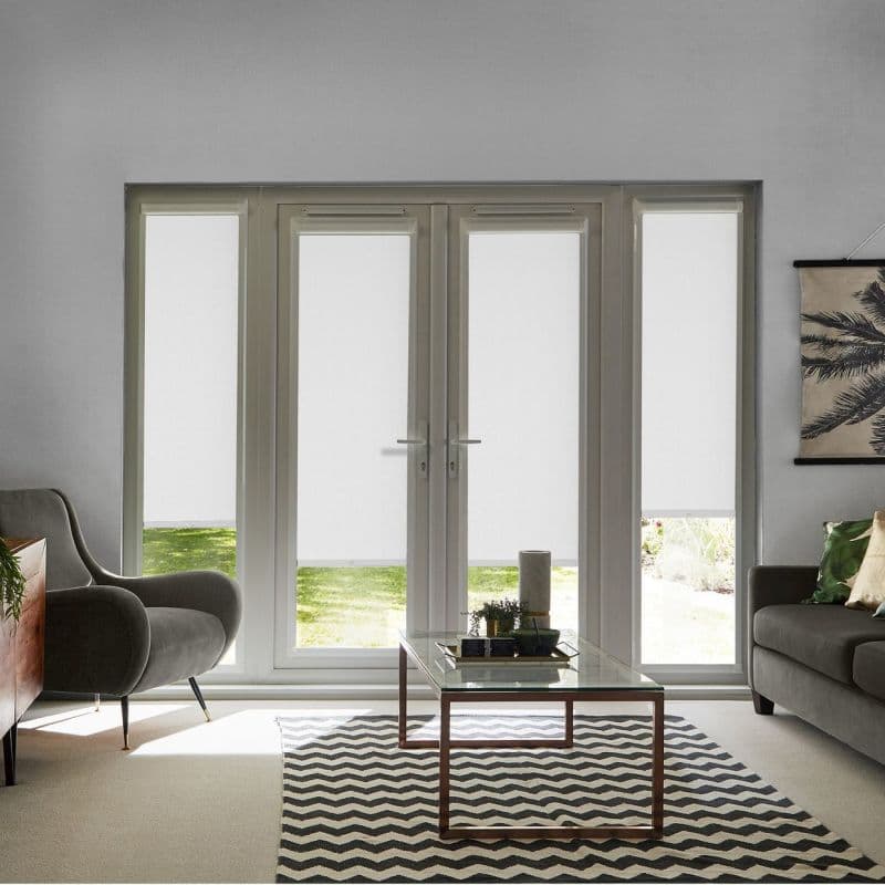 White roller blinds (inside-mounted, full-height panels) lowered across French doors and sidelights, filtering daylight with a narrow bottom reveal of lawn; modern living room with armchairs, glass coffee table, chevron rug.