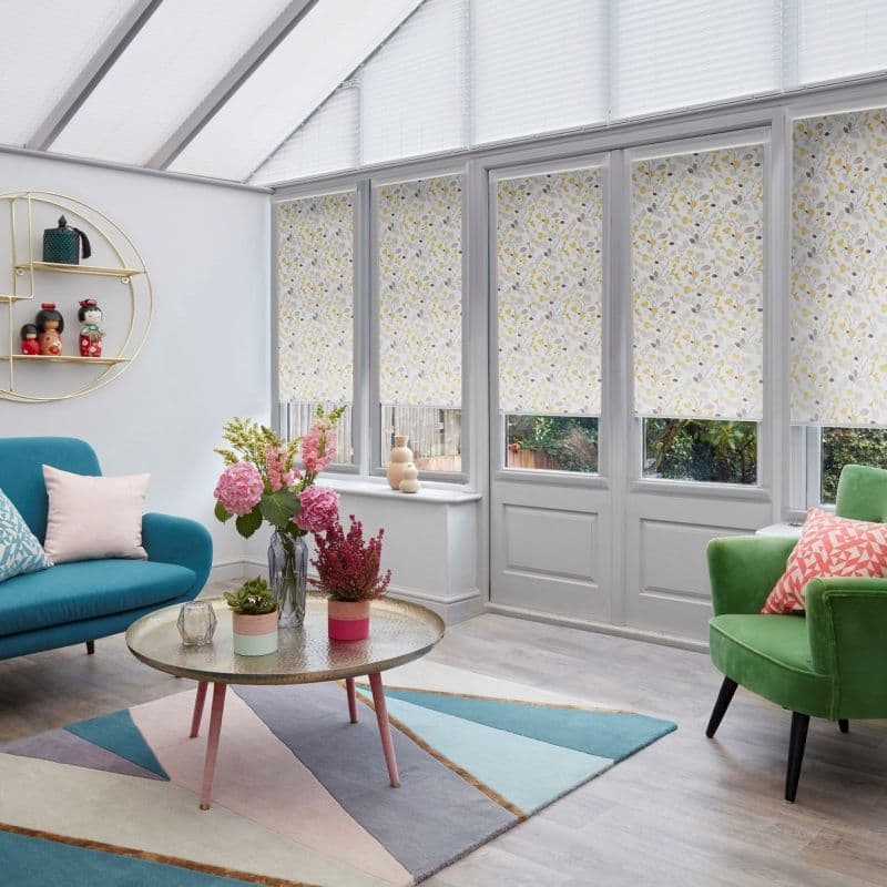 Floral-patterned roller blinds, lowered over tall conservatory windows and doors, filtering soft daylight; modern sunroom with teal sofa, green armchair, round coffee table, pastel geometric rug and potted flowers.