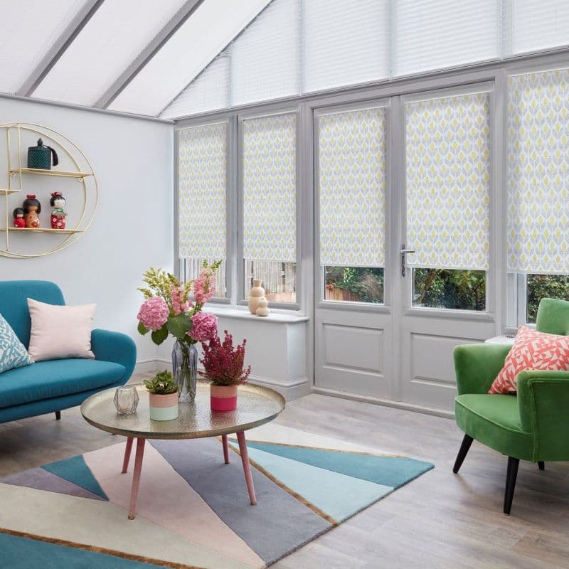 Patterned roller blinds (white with yellow-gray leaf motif) lowered across tall glazed doors and windows, filtering daylight; bright conservatory-style sitting room with teal sofa, green armchair and round coffee table.