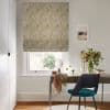 A patterned yellow-and-white Roman blind, lowered into soft folds, filtering daylight through the window; above a wooden desk with teal chair, vase of flowers, scarf and mirror in a bright room.