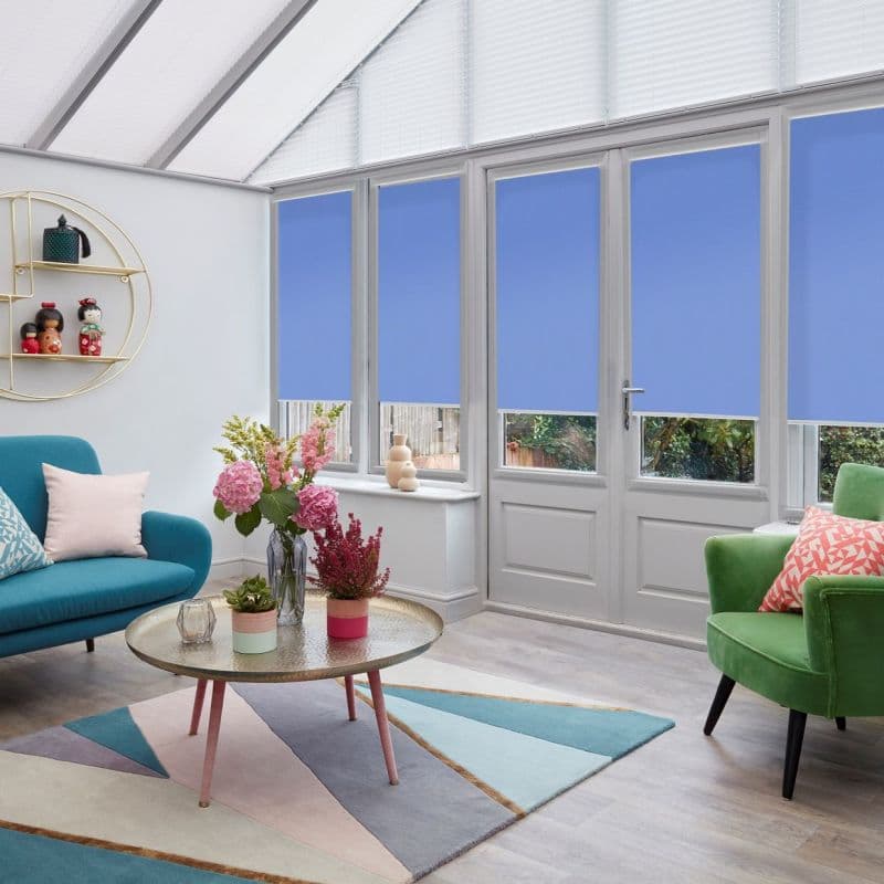 Blue roller blinds across french windows and door, lowered to cover most glass; blocking light partly, leaving narrow lower view panels — bright conservatory living room with sofas and plants.