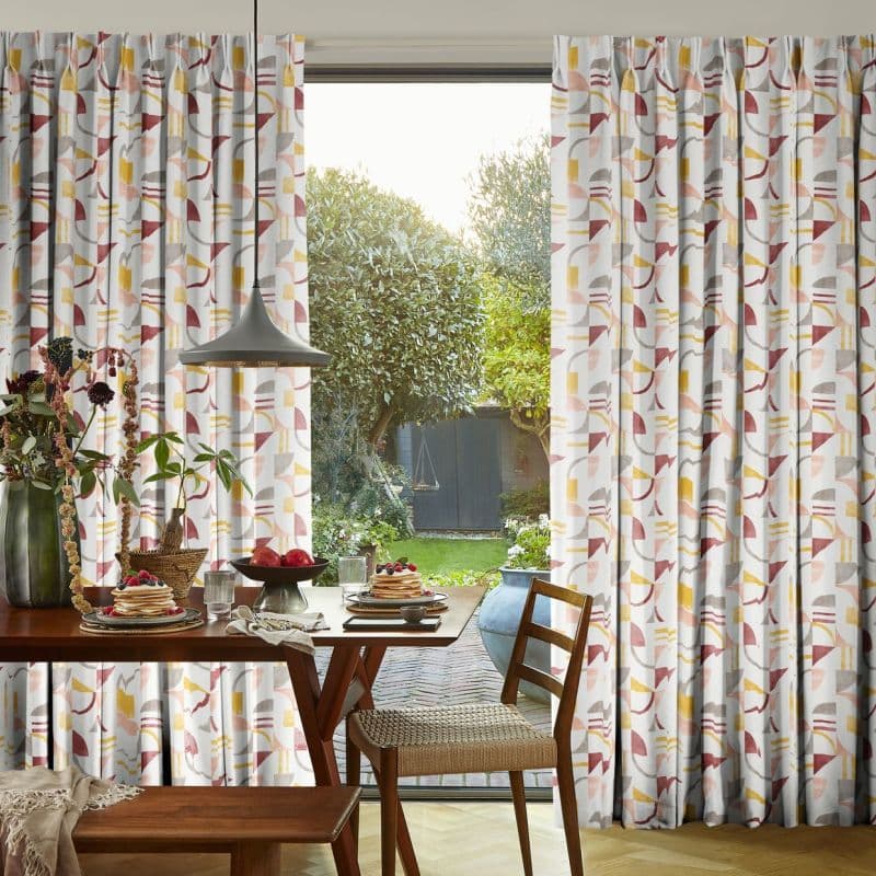 Pleated floor-length patterned curtains (pink, mustard, grey geometric print) pulled to sides, framing a glass door; filtering soft morning light over a breakfast table, pendant lamp, and garden view.