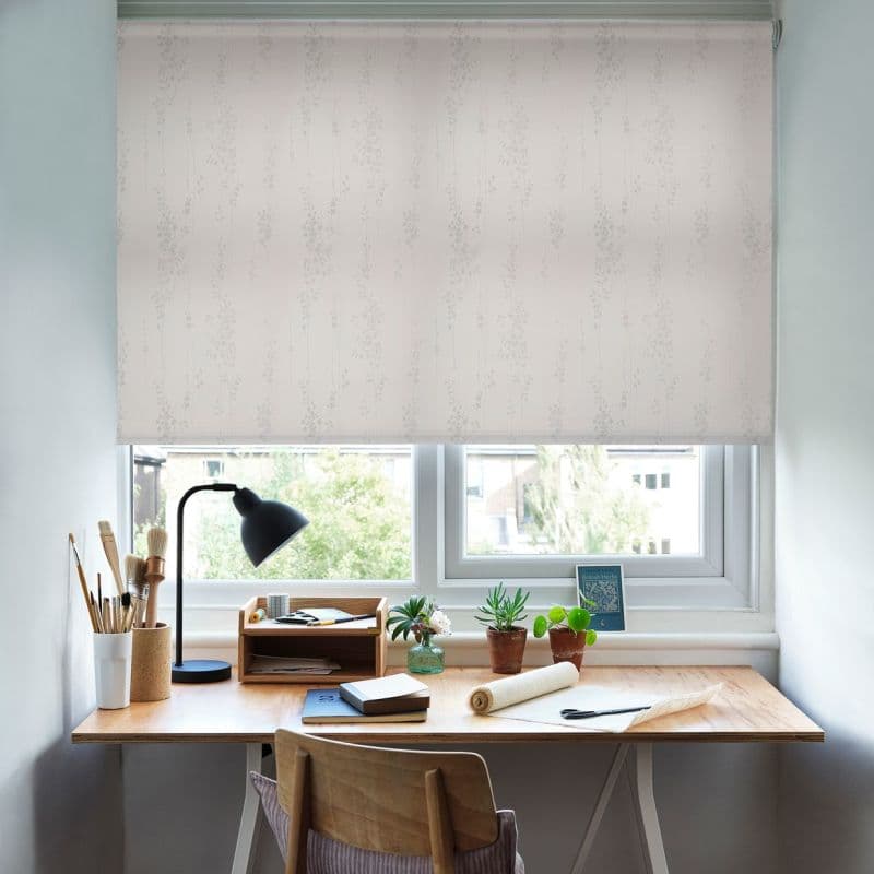 White patterned roller blind lowered across a twin window, faint vertical floral motif, filtering soft daylight into a tidy home workspace with wooden desk, lamp, brushes, potted plants, suburban view.