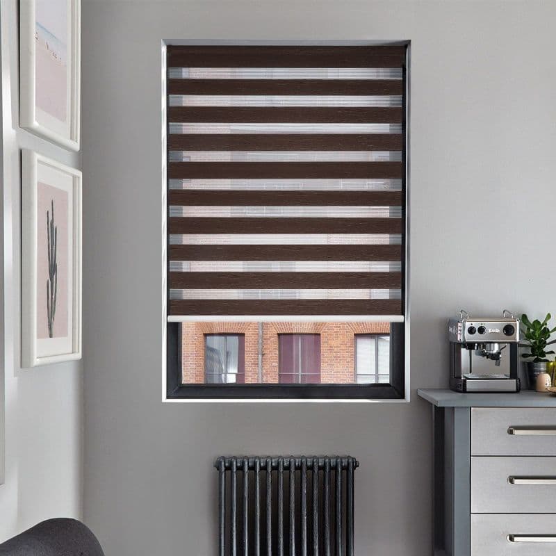 Brown zebra roller blind, lowered with alternating opaque and sheer horizontal stripes, bottom raised; filtering daylight and revealing a brick-building view in a gray kitchen with radiator and coffee machine.