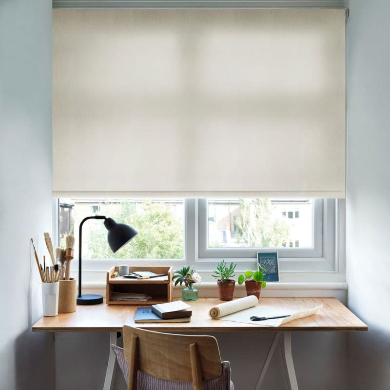 Cream fabric roller blind lowered to cover the upper window, softly diffusing daylight; overlooks a leafy suburban view above a tidy wooden desk with lamp, plants, notebooks and art brushes.