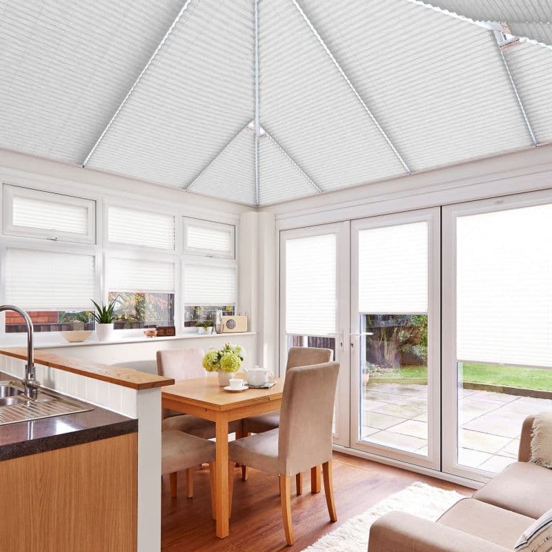 White pleated cellular blinds covering the pitched conservatory roof, windows and patio doors, closed and diffusing daylight; bright dining area with wooden table, upholstered chairs, sink and garden view.