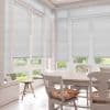 White pleated blinds covering tall French windows and patio doors, lowered to mid-height and filtering daylight; bright dining/sunroom with wooden table, chairs with floral cushions, tiled floor, garden view.
