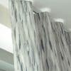 Patterned wave-fold curtains, grey-blue vertical brushstroke fabric, hanging from a ceiling-mounted track; gently pleated and drawn, filtering soft daylight in a bright, modern room near a window.