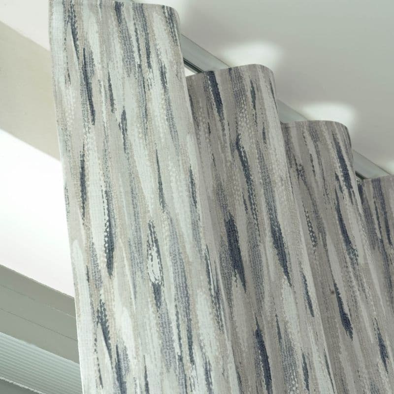 Patterned wave-fold curtains, grey-blue vertical brushstroke fabric, hanging from a ceiling-mounted track; gently pleated and drawn, filtering soft daylight in a bright, modern room near a window.