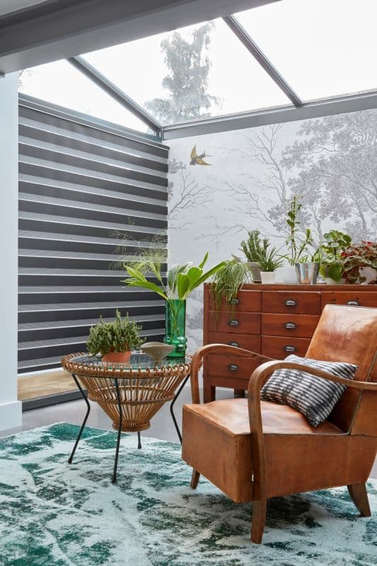 Striped dual-layer roller blind covering left glass wall, alternating dark and light horizontal bands, partially closed filtering daylight; sunroom with glass ceiling, leather armchair, rattan table, plants and wooden chest.