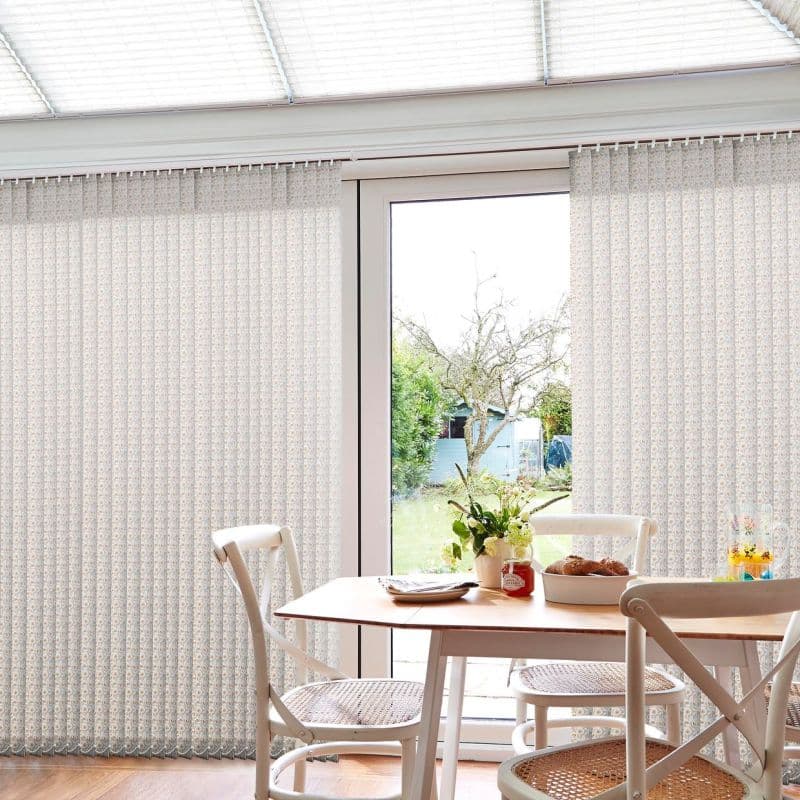 Vertical fabric blinds with pale floral pattern, spanning sliding glass doors and partially drawn, filtering soft daylight into a bright dining nook with wooden table, chairs, and a garden view.