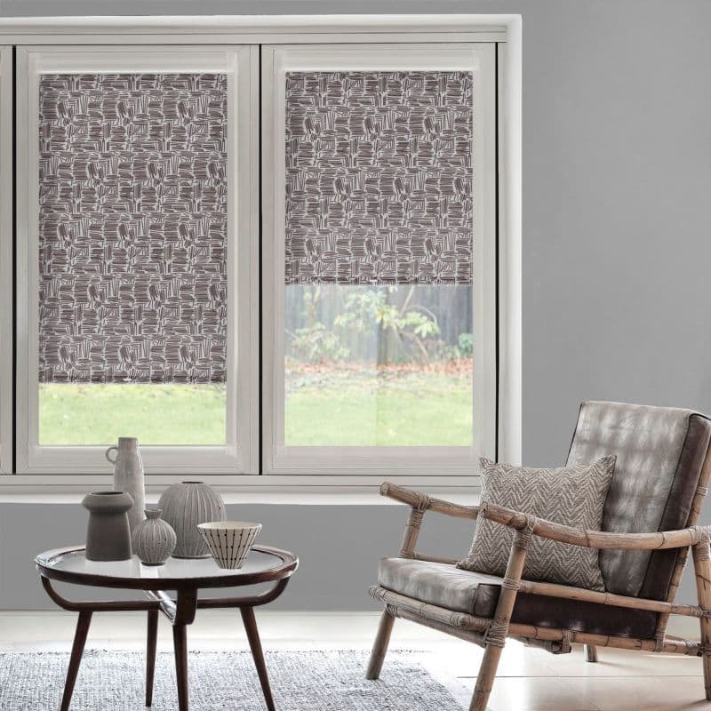 Patterned roller blinds (brown-and-white geometric) lowered halfway over two windows, partially filtering daylight; grey living room with a rattan leather armchair, coffee table, and grassy backyard view.