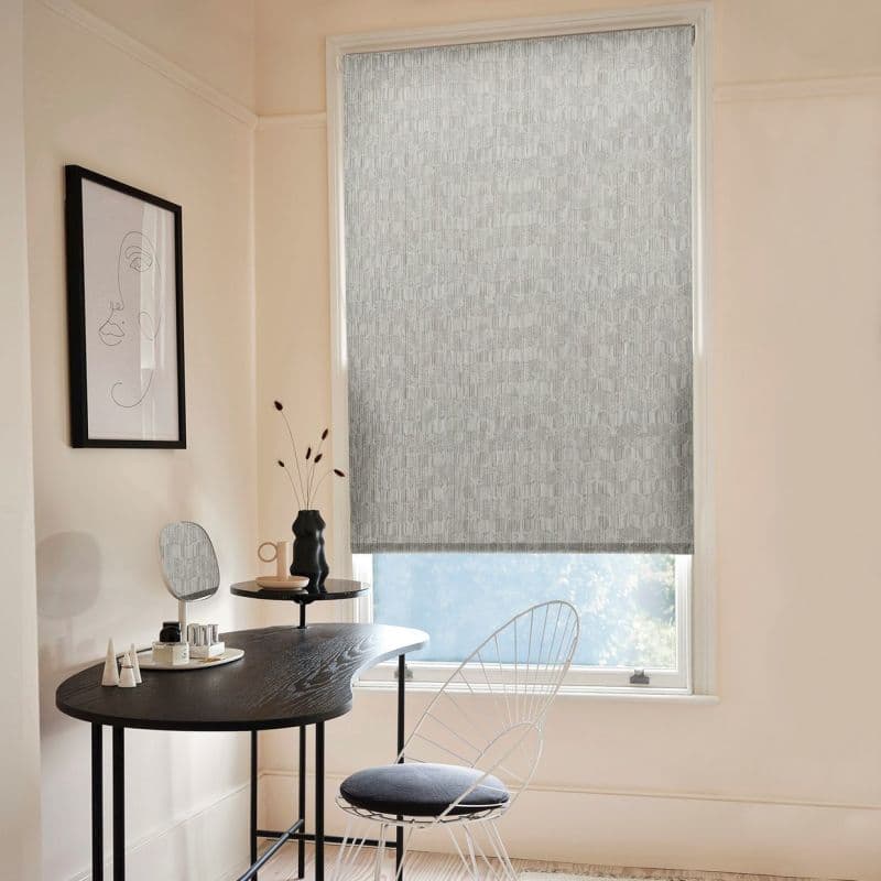 A grey patterned roller blind, lowered over a tall window, filtering daylight — in a pale minimalist dressing nook with a black desk, wire chair and framed line-art.