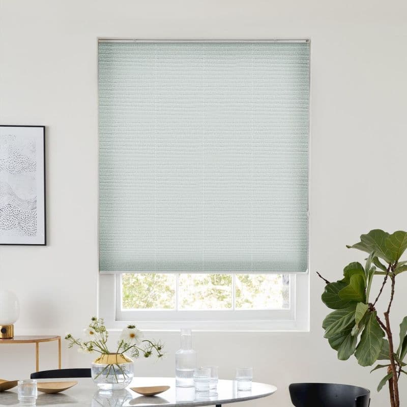 A pale green textured cellular blind, lowered over a window and filtering daylight, in a bright dining nook with round table, vase of flowers, framed art and a fiddle‑leaf fig.