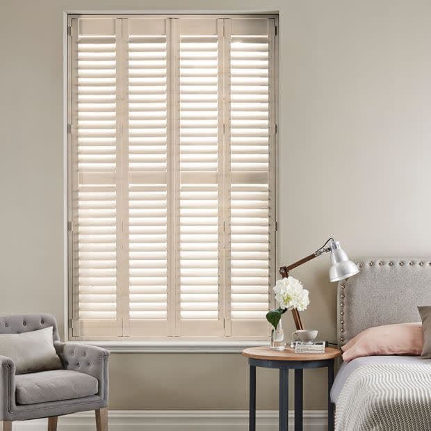 White wooden plantation shutters covering a tall inset window, closed with slatted louvers filtering soft daylight; bedroom setting with upholstered bed, bedside table, lamp and armchair in neutral tones.