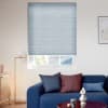 Light-blue pleated cellular blind lowered over the window, horizontally ribbed and filtering diffuse daylight; modern living room with a blue sofa, colored cushions, floor lamp, table, and rooftop/foliage view.