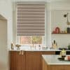 Beige horizontal pleated roller blind lowered over window, blocking most light with narrow stripes; modern kitchen sink area with brass faucet, wooden cabinets, marble countertops, island and shelving.