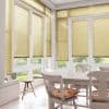 Beige pleated Venetian-style blinds cover tall French windows and doors, lowered and filtering soft daylight; in a bright tiled dining area with white table, chairs, floral cushions and garden view.