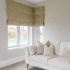 Gold leaf-patterned Roman blinds lowered across corner windows with tasselled edges, filtering daylight and covering upper panes; neutral-toned living room corner with a cream sofa, cushions, and grassy outdoor view.