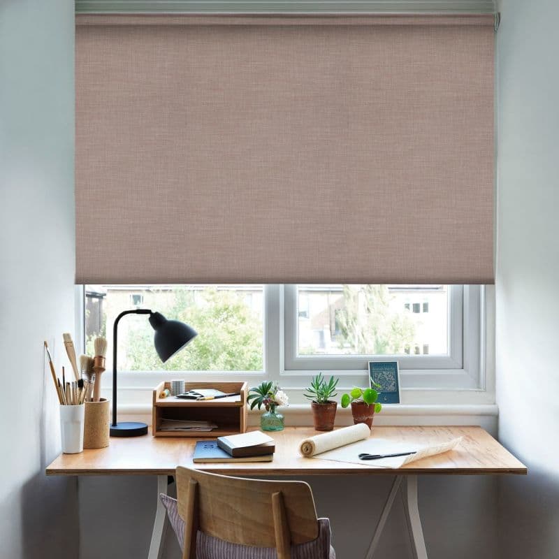 A textured, pale pink fabric roller blind lowered over a double window, filtering soft daylight; beneath it a wooden home-study desk with lamp, brushes, notebooks, small potted plants and chair.