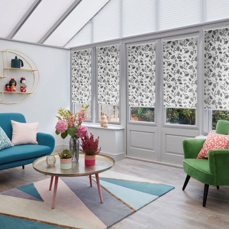 Floral-patterned roller blinds across tall conservatory windows and door, lowered to cover most glass, filtering soft daylight; surrounding bright living room with teal sofa, green armchair, and geometric rug.