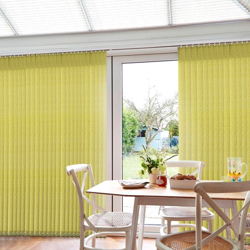 Vertical yellow patterned blinds covering a sliding glass door, parted centrally to reveal outdoors; they filter light. Dining table with white chairs and breakfast items, backyard garden with tree and shed.