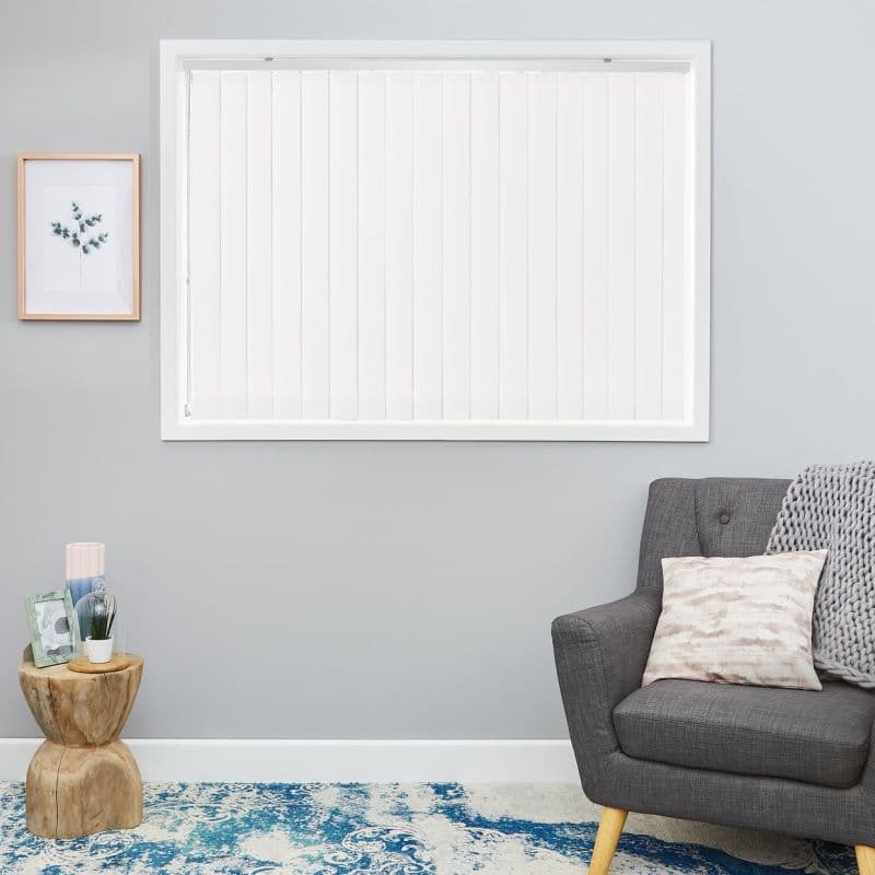 White vertical blinds covering a central window, narrow slats with cord at left; closed, diffusing daylight in a grey living room with a charcoal armchair, side table and blue-patterned rug.