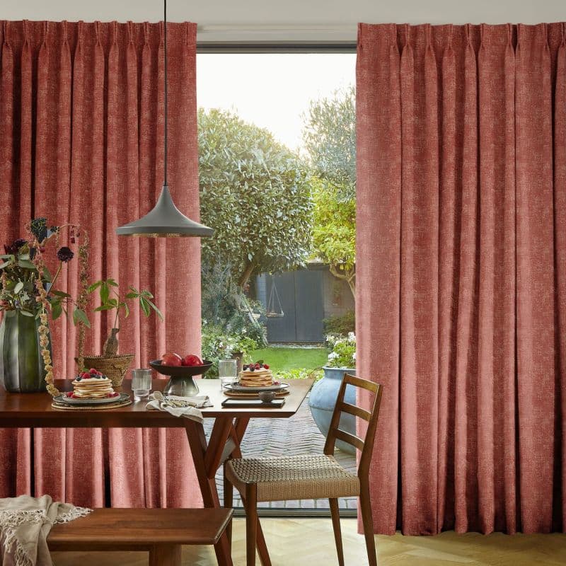Floor-length pleated textured rose curtains, parted centrally to reveal a glass door; filtering warm daylight into a dining area with wooden table, hanging pendant light, chair, and garden view.