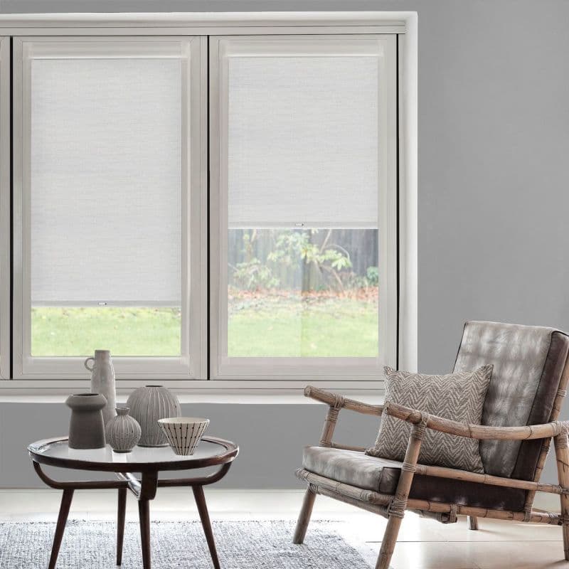 Semi-opaque white roller blinds covering twin windows, partially lowered, filtering soft daylight into a gray living room with rattan armchair, small coffee table and grassy garden view.