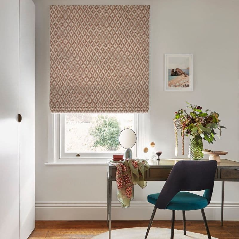 Patterned beige geometric Roman blind pulled down over sash window, softly filtering daylight. Home desk beneath holds mirror, vase of flowers and scarf; teal-upholstered chair in bright study corner.