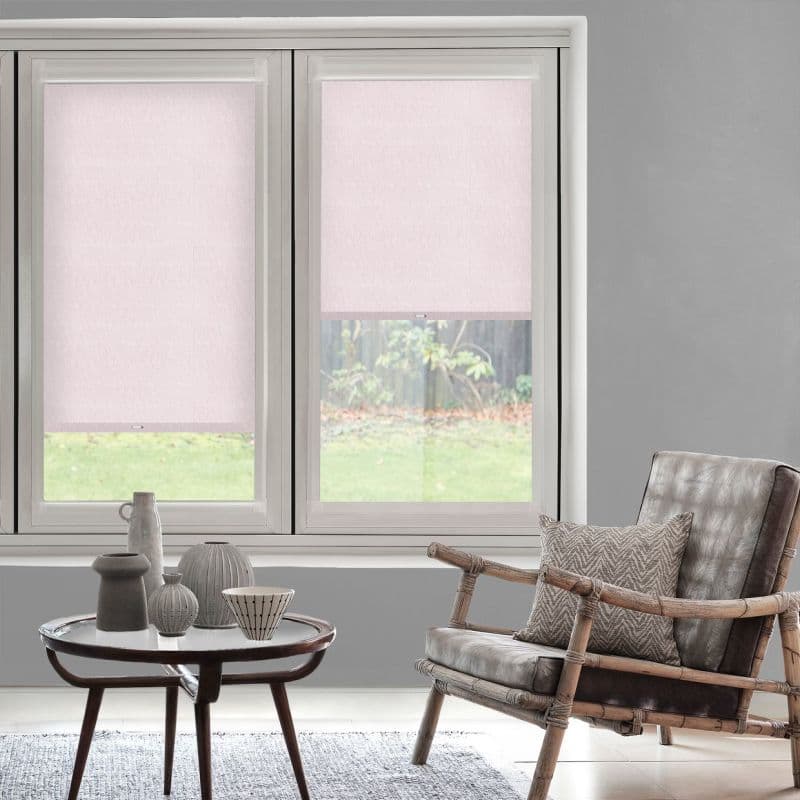 Two pale-pink fabric roller blinds covering twin windows; left fully lowered, right half-raised, gently filtering daylight onto gray-walled living room with rattan armchair, side table and backyard lawn view.