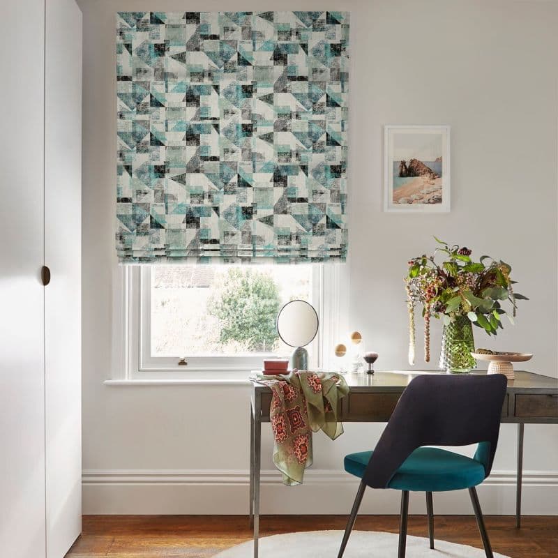 Roman blind with geometric teal-and-grey print lowered over the window, filtering daylight; desk beneath holds a mirror, vase of foliage and a teal chair in a home office.