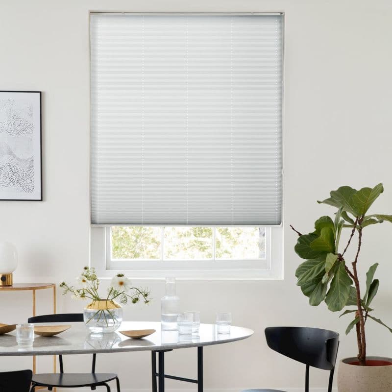 White pleated blind lowered over a recessed window, filtering soft daylight; dining table with glassware and vase, black chairs and a fiddle‑leaf fig plant in a bright minimalist room.