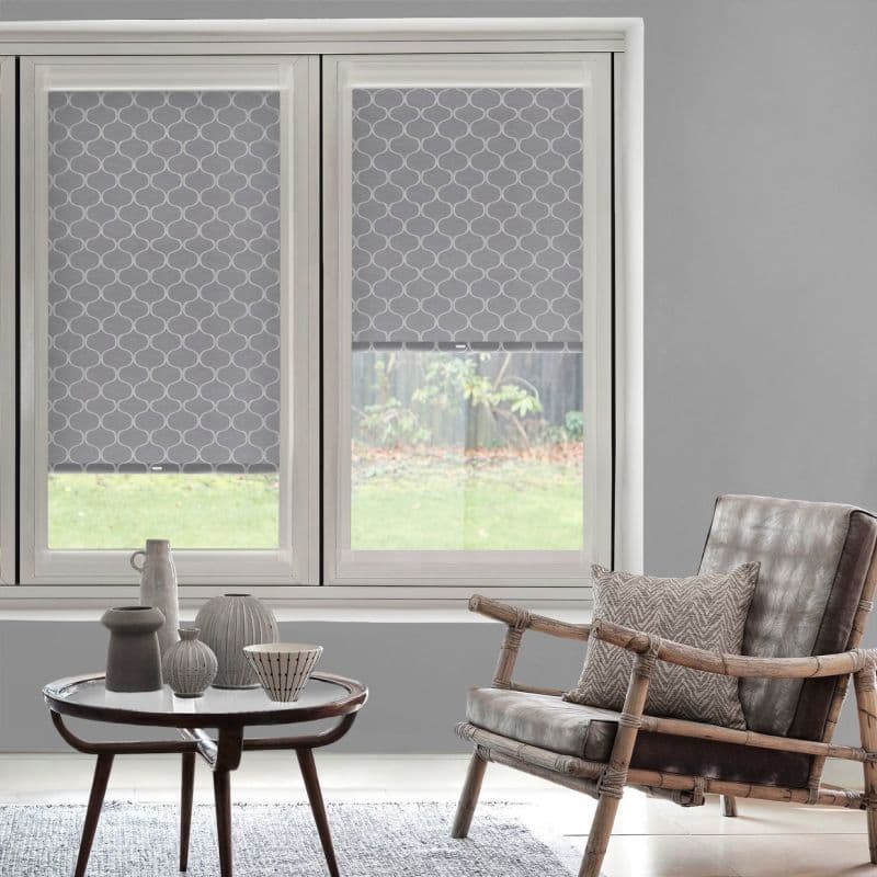 Patterned grey roller blinds with white trellis motif, inside-mounted and lowered partially, filtering daylight through the window; living room with grey walls, bamboo chair, cushion and small table with ceramics.