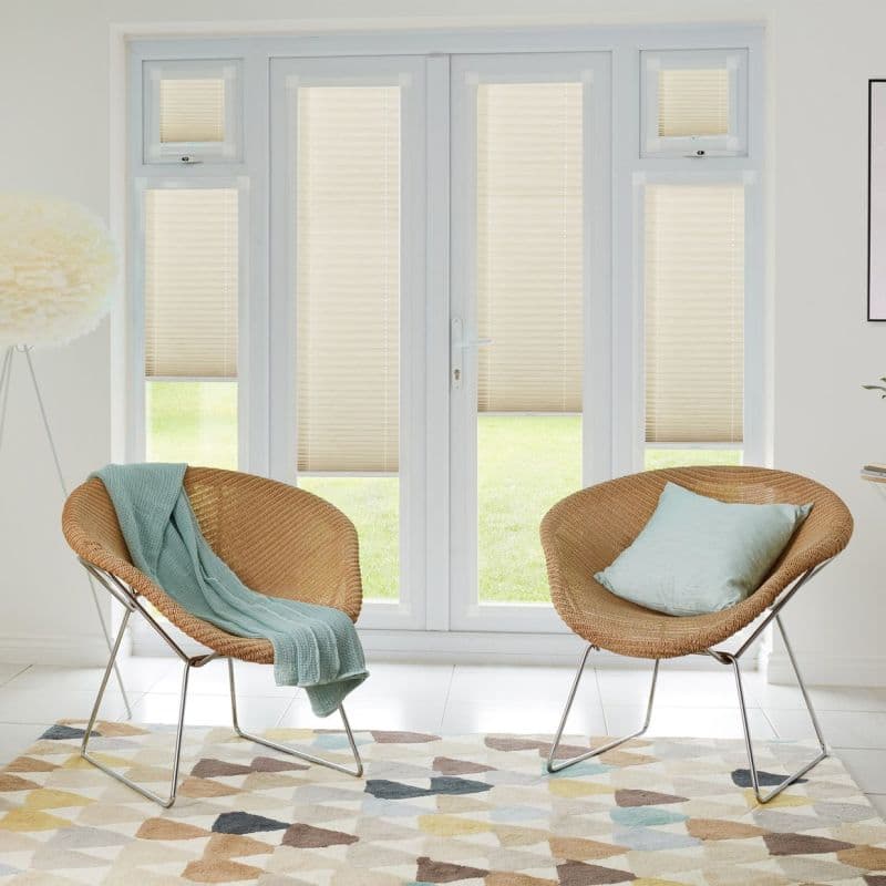 Beige pleated blinds on French doors and flanking windows, lowered to cover upper sections leaving clear lower panels, filtering daylight in living room with two wicker chairs and lawn view.