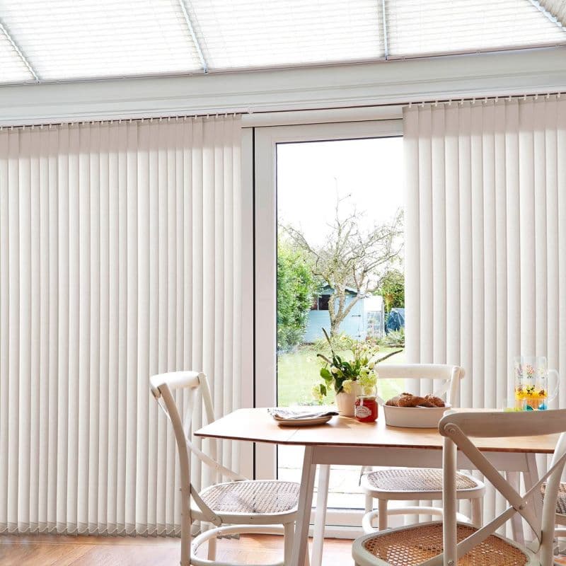 Cream vertical blinds covering patio doors, parted centrally to reveal a glass door; they hang in straight slats, softly filtering daylight in a bright dining nook overlooking a garden.
