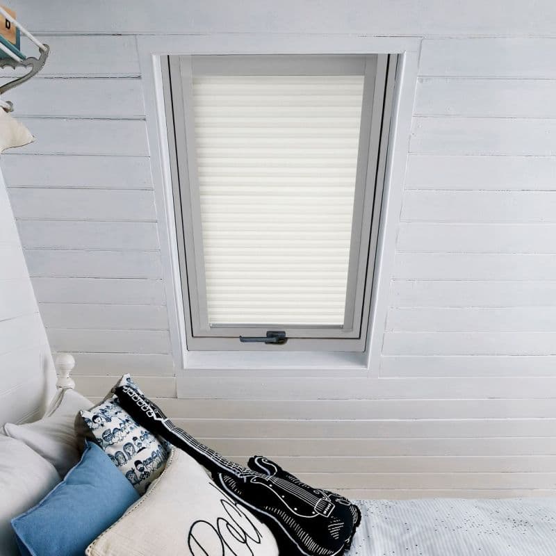 White pleated skylight blind, fully lowered with horizontal folds, filtering soft daylight; set in a white-painted attic with slatted ceiling above a bed with blue, patterned, and guitar-shaped pillows.