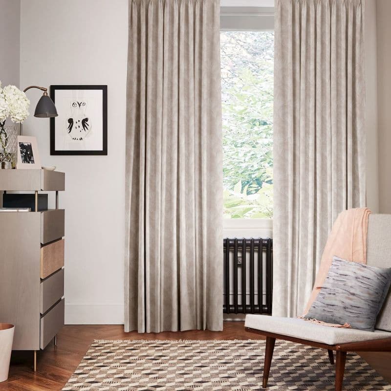 Beige patterned pleated curtains, hung floor-to-ceiling and drawn closed at sides leaving central pane exposed, filtering soft daylight; living-room setting with radiator beneath window, armchair, sideboard and patterned rug.