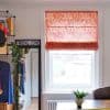 Red-orange Roman blind with white abstract-line pattern, partially lowered and filtering daylight over a sash window; bright room with radiator, desk and potted plant, wire shelving and mirror labeled FRIEND ZONE.