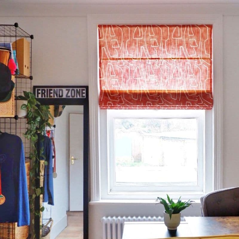 Red-orange Roman blind with white abstract-line pattern, partially lowered and filtering daylight over a sash window; bright room with radiator, desk and potted plant, wire shelving and mirror labeled FRIEND ZONE.