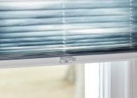 Blue pleated cellular blind, lowered with visible bottom rail and central clear plastic latch — filtering soft daylight — interior window with a blurred pale outdoor view.