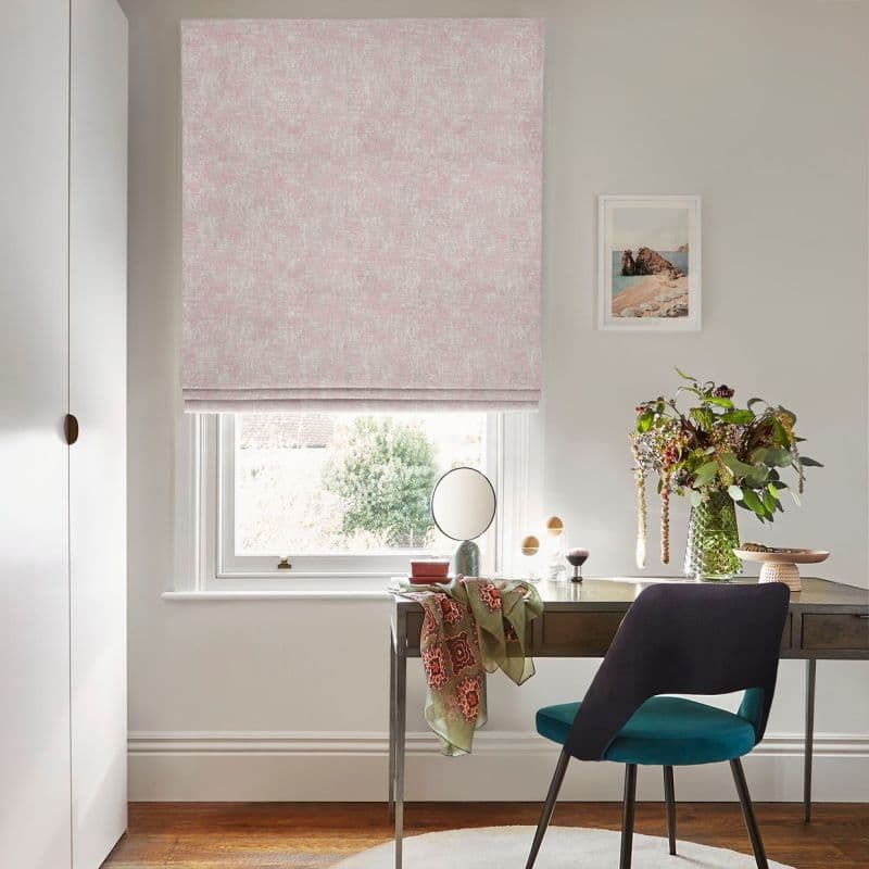 A pink textured Roman blind, fully lowered with folds, filtering daylight through a sash window above a wood desk, chair, vase of flowers and art print in a sunlit study.