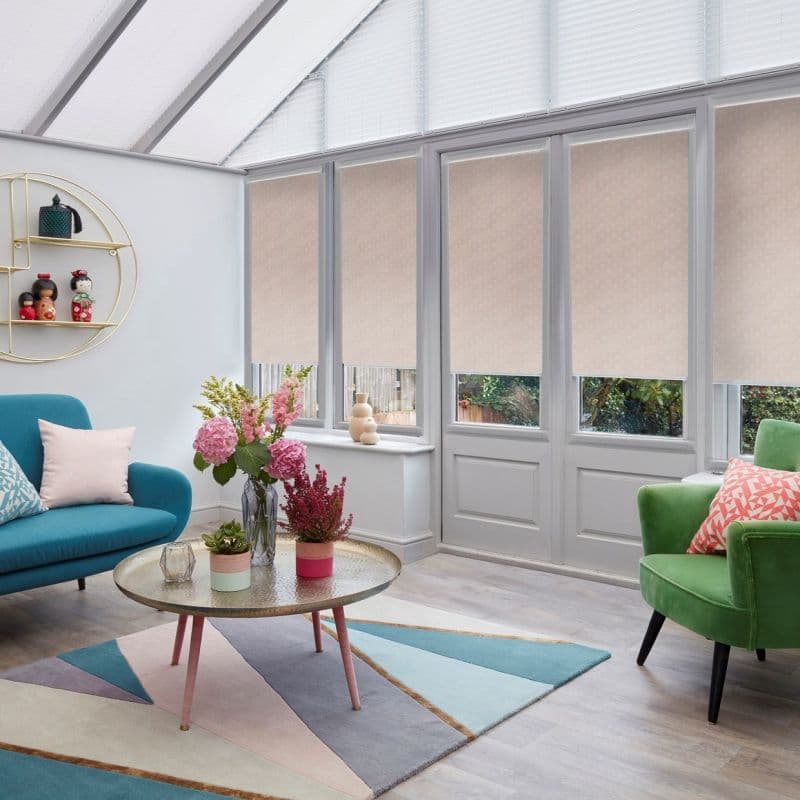 Beige roller blinds covering tall French windows and white pleated skylight blinds — lowered, filtering daylight; modern conservatory-style living room with pastel sofa, green armchair, round coffee table and flowers.
