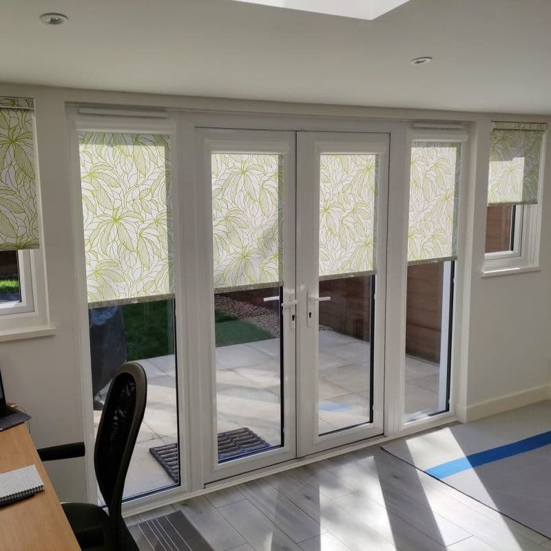 Leaf-patterned white-green roller blinds, partially lowered over four-panel French doors and side windows, filtering sunlight; small bright home office with grey tile floor and glass doors onto a patio.