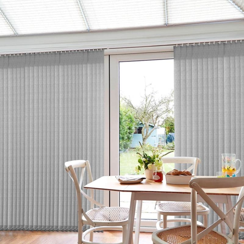 Grey vertical fabric blinds spanning patio doors, drawn closed on either side with central panel open; filtering daylight. Dining table and cane-seat chairs in bright kitchen overlooking a small garden.