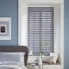 Grey plantation-style shutters (slatted panels) closed, filtering light and casting striped shadows; set in a white recessed window with a cushioned seat; pale-blue bedroom with bed, small table, vase.